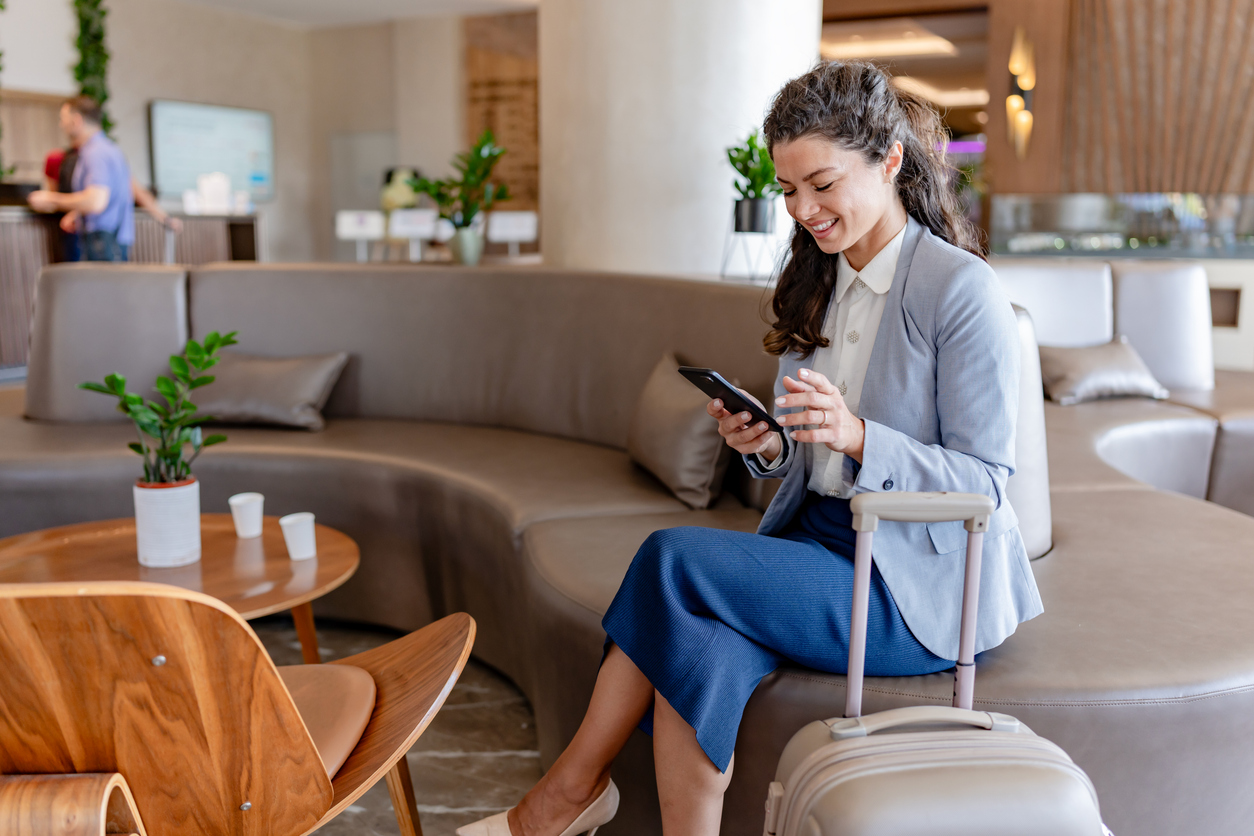 A smiling businesswoman sits in a modern hotel lobby, checking her phone while holding onto her suitcase. She appears relaxed and ready for her trip, surrounded by a comfortable lounge area